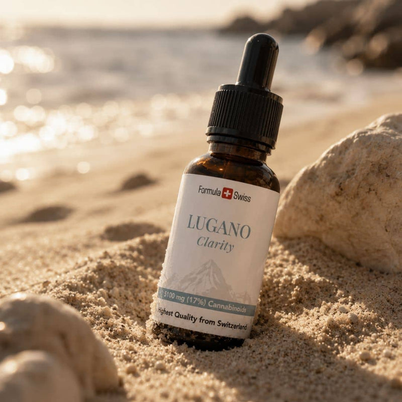 Formula Swiss Lugano Clarity CBD olja i en flaska, placerad i strandsand bredvid stenar och solbelyst vatten.